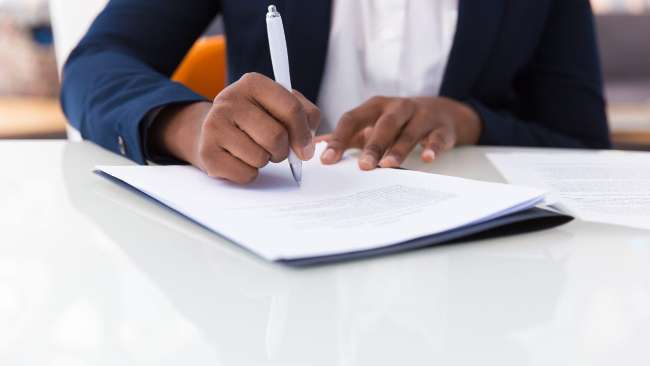 Businesswoman signing contract. African American business woman sitting at table in office, holding pen and writing in document. Legal expertise concept